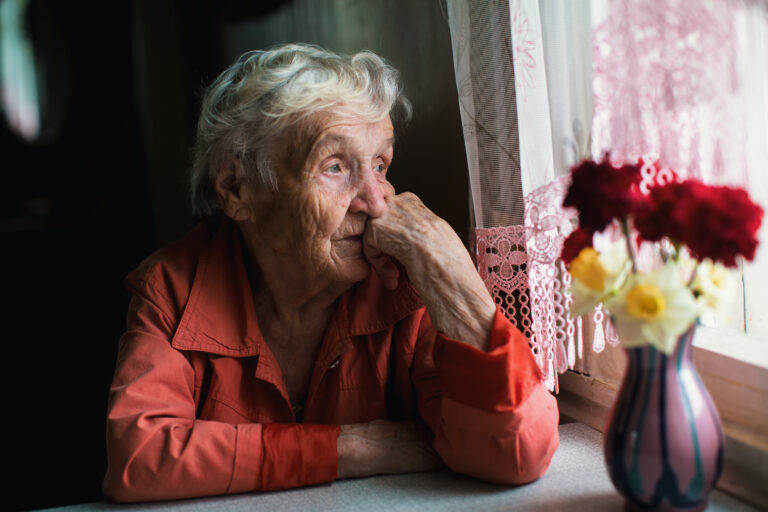 Eldre kvinne som sitter ved kjøkkenbordet og ser ut gjennom vinduet